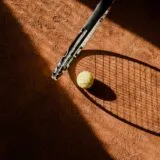 Rachetă de tenis și câteva mingi galbene pe un teren de zgură, lângă linia de fund