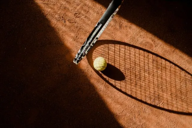 Rachetă de tenis și câteva mingi galbene pe un teren de zgură, lângă linia de fund